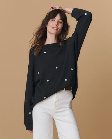 A woman wearing THE GREAT. PF25 EMB KNITS The Slouch Sweatshirt, embroidered in almost black with daisy designs, paired with white pants, stands against a beige background.