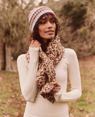Standing outdoors with trees in the background, a person is wearing a colorful knitted hat, a white long-sleeve top, and *The Down Quilted Puffer Scarf* in Snow Leopard by THE GREAT. FALL 24 TGO, completing their stylish look.