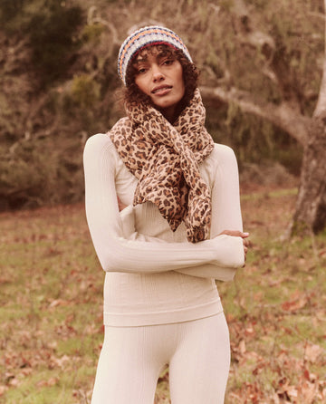 An individual in a beige outfit and leopard-print scarf stands in a grassy area with trees in the background, wearing The Down Quilted Puffer Scarf in Snow Leopard by THE GREAT. FALL 24 TGO, ensuring warmth and style with its unique down puffer accessories.