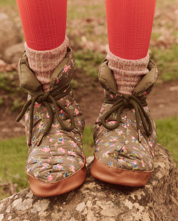 A person stands on a rock wearing The Down Quilted Puffer Slipper in Cypress Basin Floral by THE GREAT. FALL 24 TGO, paired with pink socks and red leggings, highlighting their flair for technical clothing with brown soles.