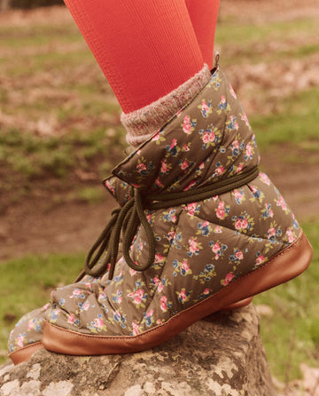 A person in The Down Quilted Puffer Slipper in the Cypress Basin Floral pattern from THE GREAT. FALL 24 TGO, paired with red leggings and gray socks, standing on a rock.