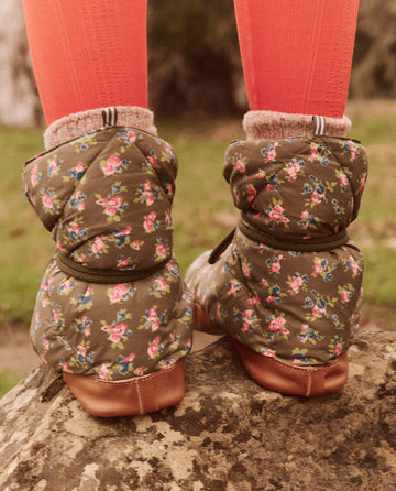 A person wearing The Down Quilted Puffer Slipper in the Cypress Basin Floral design from THE GREAT. FALL 24 TGO, paired with orange socks, stands on a rock outdoors, highlighting clothing committed to the Responsible Down Standard.