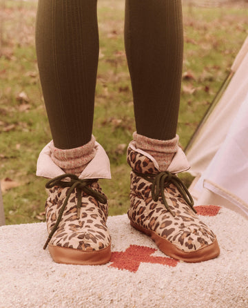 A person wearing The Down Quilted Puffer Slipper in Snow Leopard from THE GREAT. FALL 24 TGO and green leggings, standing on a textured surface with grass in the background, accessorized with a stylish down puffer.