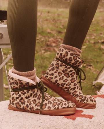 A person wearing The Down Quilted Puffer Slipper in Snow Leopard and green leggings stands on a rug outdoors, embracing the spirit of technical clothing with their bold fashion choice from THE GREAT. FALL 24 TGO collection.