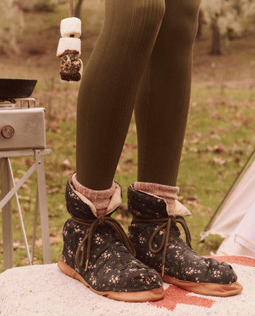 The individual stands on a blanket, wearing Wilderness Floral boots and green leggings, while holding a skewer with toasted marshmallows. Nearby, The Down Quilted Puffer Slipper in Powder Pink from THE GREAT.'s FALL 24 TGO collection lies beside the camping stove, enhancing the cozy atmosphere against the grassy backdrop.