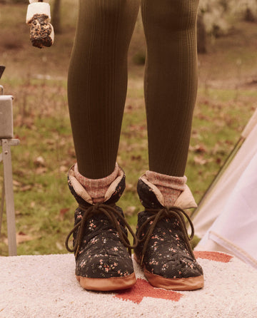 A person stands on a rug next to a tent, wearing boots with a floral pattern paired with green tights, and accessorized with visible socks and The Down Quilted Puffer Slipper in Wilderness Floral and Powder Pink by THE GREAT. FALL 24 TGO.