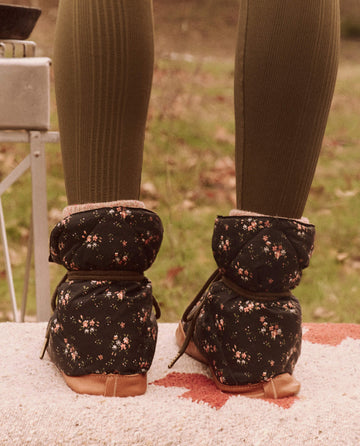 A person wearing Wilderness Floral and Powder Pink Down Quilted Puffer Slippers from THE GREAT. FALL 24 TGO stands on a textured surface, with grass in the background.