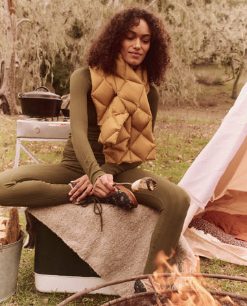 By the campfire, someone sits cross-legged on a cooler, roasting a marshmallow. They're wearing a green outfit paired with The Down Quilted Puffer Scarf in Goldenrod from THE GREAT. FALL 24 TGO collection, which is ideal for outdoor adventures thanks to its waterproof and breathable fabrics. In the background, a pot simmers on the portable stove.