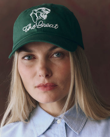 A woman in a green THE GREAT. Baseball Hat with vintage panther embroidery and "The Great" text, paired with a light blue collared shirt, looks directly at the camera.