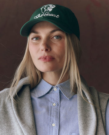 A woman with long blonde hair and a neutral expression wears THE GREAT. F25 D1 ACCESSORIES The Baseball Hat. Graphic -- Green, featuring vintage "The Great" and bear embroidery, paired with a light blue button-up shirt and gray sweater.