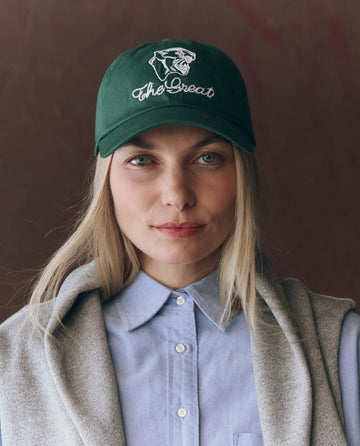 A woman with straight blonde hair wears THE GREAT. F25 D1 ACCESSORIES "The Baseball Hat. Graphic -- Green," featuring vintage panther embroidery, paired with a light blue button-up shirt and a gray sweater over her shoulders.