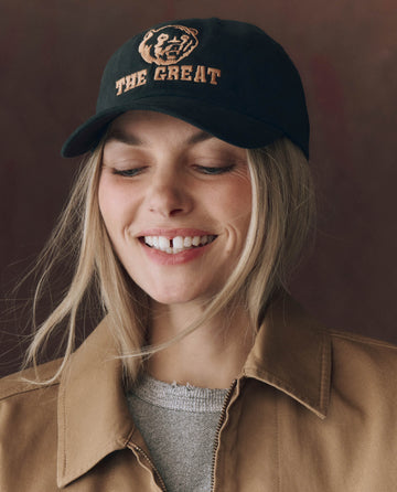 A woman wearing THE GREAT. F25 D1 ACCESSORIES The Baseball Hat. Graphic -- Black, featuring "THE GREAT" in chainstitch embroidery, smiles while looking slightly down, paired with a tan jacket and gray top.