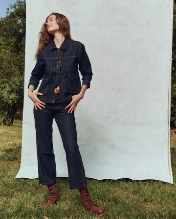 A person stands on grass, wearing THE GREAT. PS25's The Workwear Jean in Rinse and brown boots, smiling and looking to the side in front of a light backdrop.