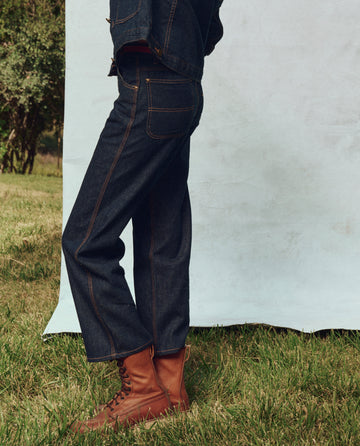 A person in The Cropped Workwear Jean by THE GREAT. PS25 stands on grass in rugged brown boots against a light blue backdrop, showcasing contemporary workwear style.