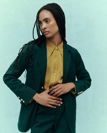 A person with long braided hair stands against a light green background, wearing The Tailored Blazer in Winter Pine and a yellow shirt from THE GREAT. HOL 24 collection.