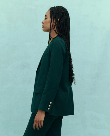 A person wearing The Tailored Blazer in Winter Pine by THE GREAT. HOL 24 and matching pants stands in profile against a light teal background.