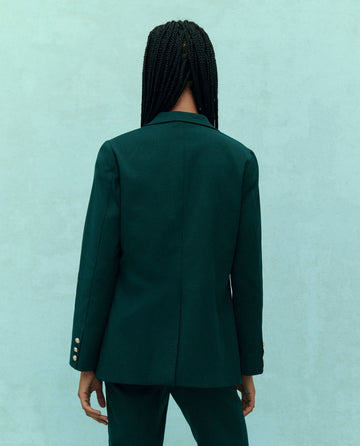 A person with long braided hair wearing The Tailored Blazer in Winter Pine from THE GREAT. HOL 24 stands facing away against a light teal background.
