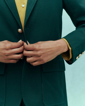 A person in The Tailored Blazer - Winter Pine fastens the jacket, revealing a yellow shirt underneath. This stylish piece is from THE GREAT.'s HOL 24 collection.