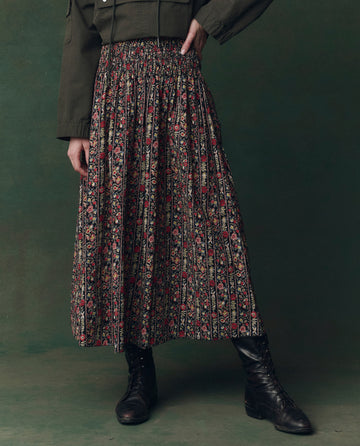 Person wearing a dark green jacket, THE GREAT. F25 D2 The Canary Skirt in Garland Print, and black lace-up boots, standing against a green background.