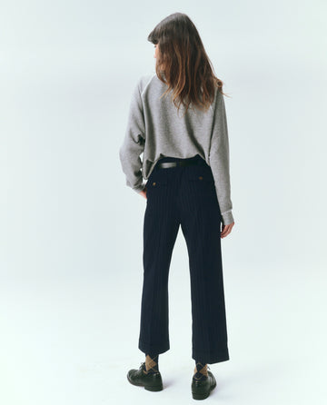 A person with long hair stands facing away, wearing The College Sweatshirt in Varsity Grey from THE GREAT. CORE KNITS, along with dark pants and black shoes. The background is plain and light-colored.