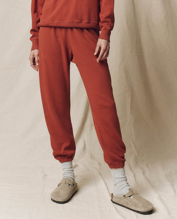 Wearing THE GREAT. F25 SEASONAL KNITS The Stadium Sweatpant in Brick, gray socks, and beige clogs, this person stands before a neutral fabric backdrop, showcasing laidback Los Angeles style.