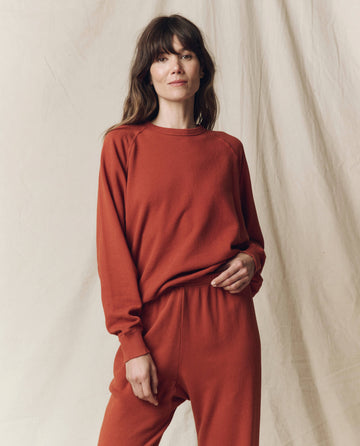 A woman stands against a beige backdrop wearing THE GREAT. F25 SEASONAL KNITS’ The College Sweatshirt. Solid -- Brick, an oversized, rust-colored set.