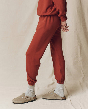 A person in THE GREAT. F25 SEASONAL KNITS The Stadium Sweatpant. Solid -- Brick stands against a neutral fabric backdrop, showcasing a relaxed Los Angeles-inspired look with matching pieces, beige socks, and taupe slip-on sandals.
