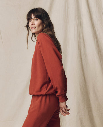A woman with long brown hair wears THE GREAT.'s The College Sweatshirt in Solid Brick from F25 SEASONAL KNITS, paired with matching pants. She stands before a beige fabric backdrop, evoking vintage inspiration and handcrafted in Los Angeles.