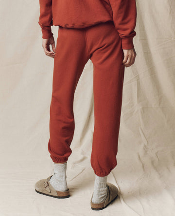 A person stands on a light fabric backdrop, shown from the waist down and facing away, wearing THE GREAT. F25 SEASONAL KNITS The Stadium Sweatpant. Solid -- Brick with a matching top, gray socks, and beige sandals. Handcrafted in Los Angeles.