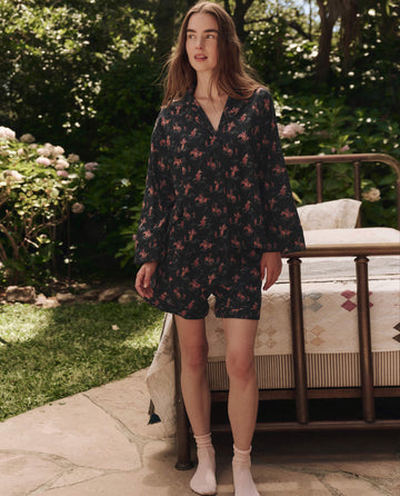 A woman stands on a stone patio near a bed, wearing THE GREAT. SP25 SLEEP's The Pajama Shirt in Navy Cowboy. Light socks and a backdrop of green plants and trees create a cozy sleepwear atmosphere.