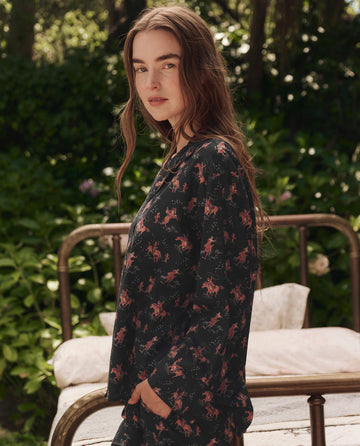 A woman with long brown hair stands outdoors in front of a brass bed, wearing THE GREAT. SP25 SLEEP's The Pajama Shirt in Navy Cowboy, and looks at the camera with lush greenery behind her.
