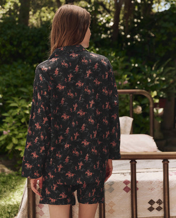 A person with long hair stands outdoors near a metal bed in a garden, wearing THE GREAT. SP25 SLEEP’s The Pajama Shirt in Navy Cowboy—coordinated dark sleepwear with a pink pattern, facing away from the camera.