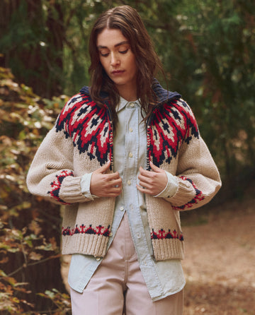 A person stands in a wooded area wearing THE GREAT. F25 TGO's The Nordic Sweater -- Glacier Fair Isle over a light blue button-up shirt and beige pants, looking down while holding the sweater.