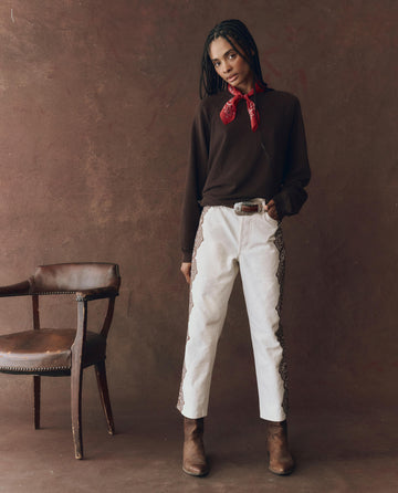 A person stands indoors in a brown sweater, brown boots, a red neck scarf, and THE GREAT. F25 D1 The Lace Print Wayne Jean in cream with brown. A wooden chair sits to their left against a brown backdrop.