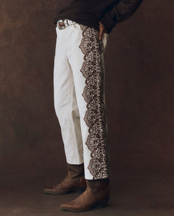 A person wearing THE GREAT. F25 D1 The Lace Print Wayne Jean in cream with brown, styled with a dark sweater, brown leather belt, and cowboy boots stands against a dark background.
