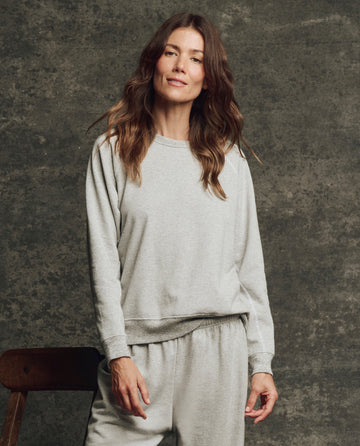 A woman with wavy brown hair wears THE GREAT. H25 CORE FLEECEBACK The Fleece Rookie Sweatshirt in heather grey, standing indoors against a textured grey background.