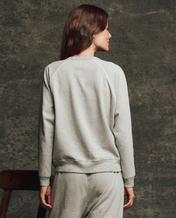 A woman with long brown hair wears THE GREAT. H25 CORE FLEECEBACK's The Fleece Rookie Sweatshirt in Heather Grey, paired with matching sweatpants, standing with her back to the camera against a dark textured background.
