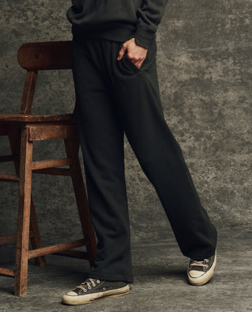 A person wearing THE GREAT. H25 CORE FLEECEBACK The Fleece Victory Sweatpant in Almost Black and black sneakers stands by a wooden chair, one hand in their pocket. Only the lower body is visible.