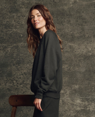 A woman with long wavy brown hair wears THE GREAT. H25 CORE FLEECEBACK "The Fleece College Sweatshirt" in Almost Black, standing sideways beside a wooden chair against a textured dark background.