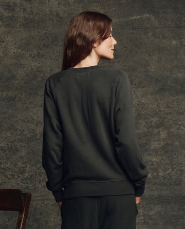 A woman with long brown hair faces away from the camera, wearing THE GREAT. H25 CORE FLEECEBACK The Fleece College Sweatshirt in Almost Black, in front of a textured gray wall.