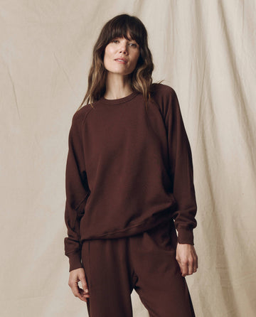 A person wears THE GREAT.'s Driftwood College Sweatshirt from the F25 SEASONAL KNITS line, paired with matching brown pants, standing before a beige fabric backdrop.