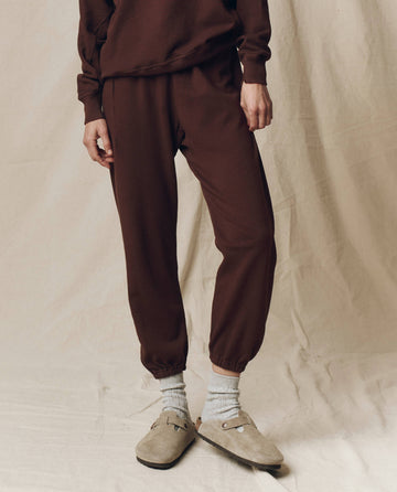 Wearing THE GREAT. F25 SEASONAL KNITS The Stadium Sweatpant, Solid -- Driftwood, paired with a matching sweatshirt, gray socks, and beige suede slip-ons on a beige fabric backdrop—embodying relaxed Los Angeles style.