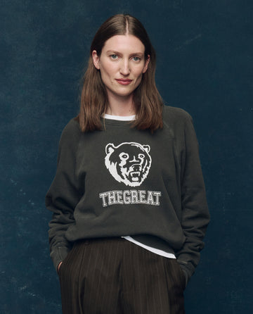 A woman with straight brown hair models THE GREAT. SP25 COLLECTORS "The College Sweatshirt" in washed black with Bear Graphic, standing against a dark blue textured background.