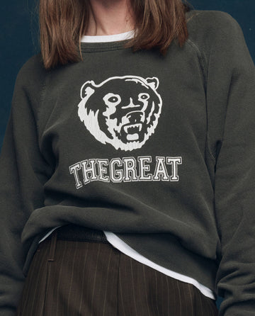 A person wearing The College Sweatshirt by THE GREAT. SP25 COLLECTORS in washed black, featuring a bear graphic and "THE GREAT" on the front.
