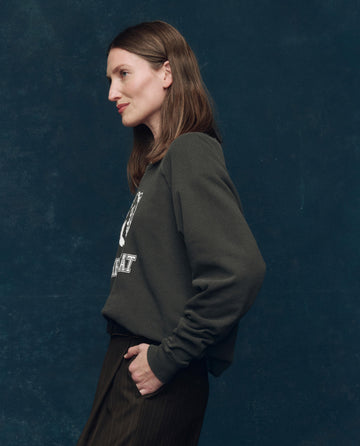 A woman with straight brown hair stands in profile against a dark blue background, wearing THE GREAT. SP25 COLLECTORS The College Sweatshirt in washed black with bear graphic and pinstriped pants, one hand tucked in her pocket.