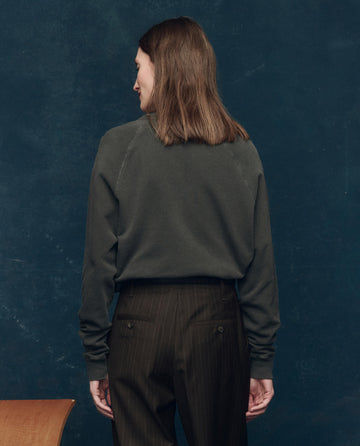 A person with shoulder-length brown hair, wearing THE GREAT. SP25 COLLECTORS' College Sweatshirt in washed black with Bear graphic and pinstriped trousers, stands facing away against a dark blue background.
