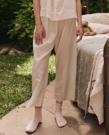 A person stands outdoors on a stone path, wearing THE GREAT. SP25 SLEEP Eyelet Easy Sleep Pant in True White, crafted from cotton voile, with pink socks and a cream top; nearby is a bed with a quilt and teddy bear.