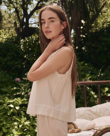 A young woman with long hair stands outdoors near a bed, wearing THE GREAT. SP25 SLEEP The Eyelet Tank in True White, surrounded by greenery and sunlight.