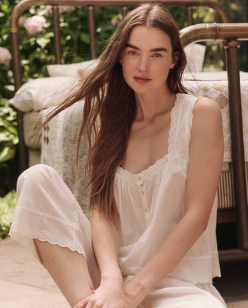 A woman with long brown hair sits outside by a metal bed, wearing THE GREAT. SP25 SLEEP’s The Eyelet Tank in True White, made of soft cotton voile, looking at the camera with a neutral expression.