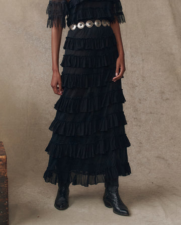 Model wears THE GREAT. H25 COLLECTION's "The Winter Mesh Flounce Skirt" in black, styled with a wide silver belt and black boots, standing against a neutral textured background.
